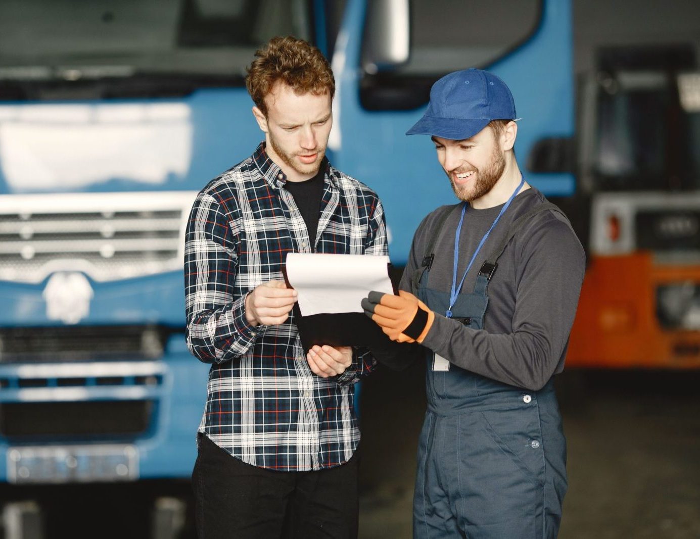 Two men in discussion over vehicle maintenance documents in a garage setting with trucks in the background.